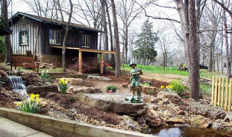 Landscaped area with a small pond, statuary, and a waterfall in front of a cabin in the woods