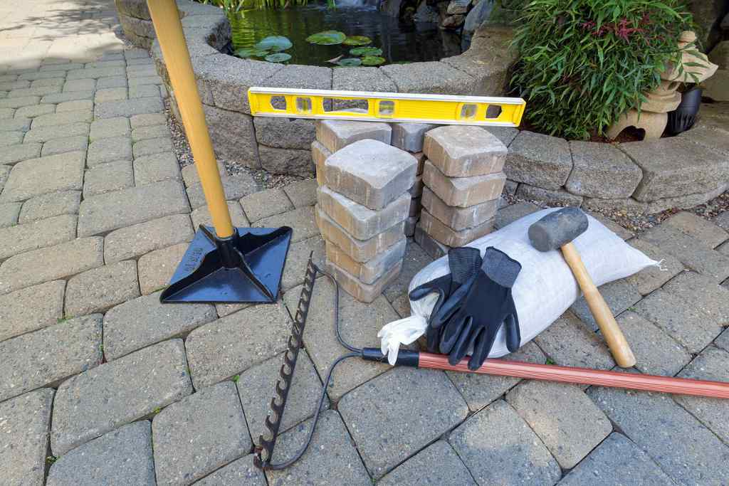 Stone Pavers for backyard patio with tools of landscaping services near me, including gloves, level, rubber mallet, and a sand and gravel tamper.