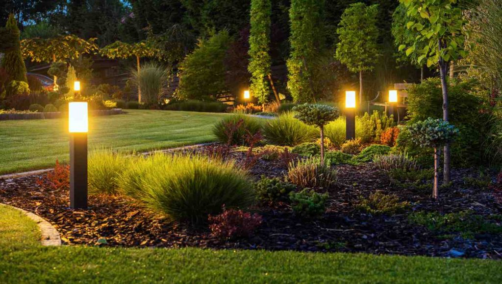 A photo of a beautifully landscaped yard in the dark, illuminated by yellow lamp posts. Done by professional landscapers and landscaping experts.