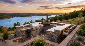 A panoramic sunset photograph of a custom stacked-stone and granite outdoor kitchen overlooking Lake of the Ozarks. The luxury setup includes a built-in grill, pizza oven, bar seating, and integrated lighting, built on a tiered patio on a sloped waterfront lot by Heartland Landscape Group.