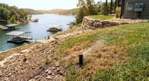 A pop-up sprinkler head spraying weakly over a dry brown patch of grass on a steep, rocky sloped lawn overlooking Lake of the Ozarks, with boat docks and a smart controller visible in the background.